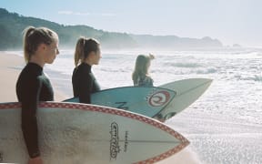 The promotional poster for Over the Undertow. Three women stand side by side on the beach, wearing wetsuits and holding surfboards. They look out at the ocean.