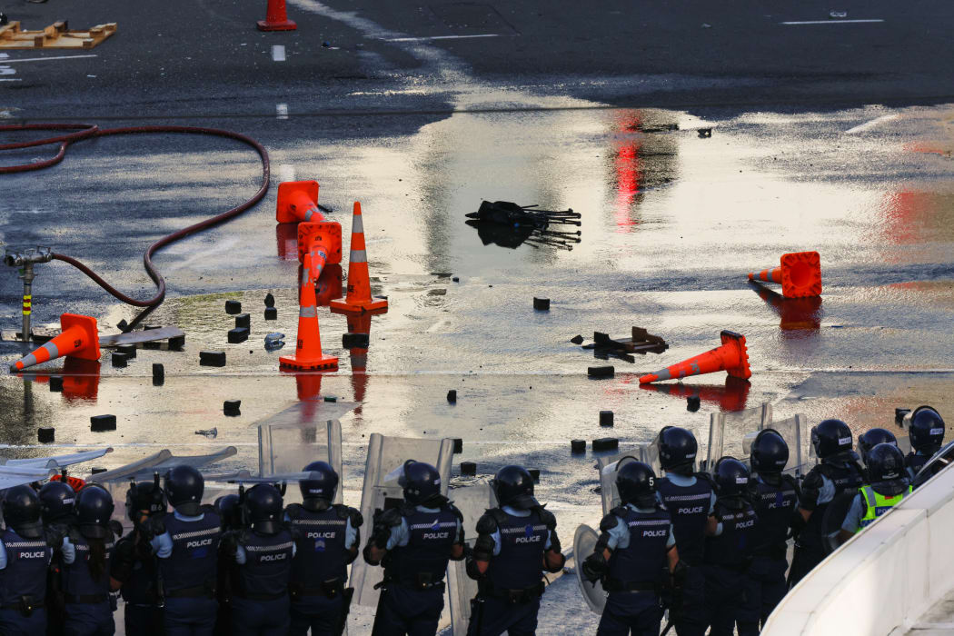 A retreated police line stands silhouetted against the corner of Lambton Quay and Bowen Street, littered with bricks and cones that have been thrown at them in an earlier position.
