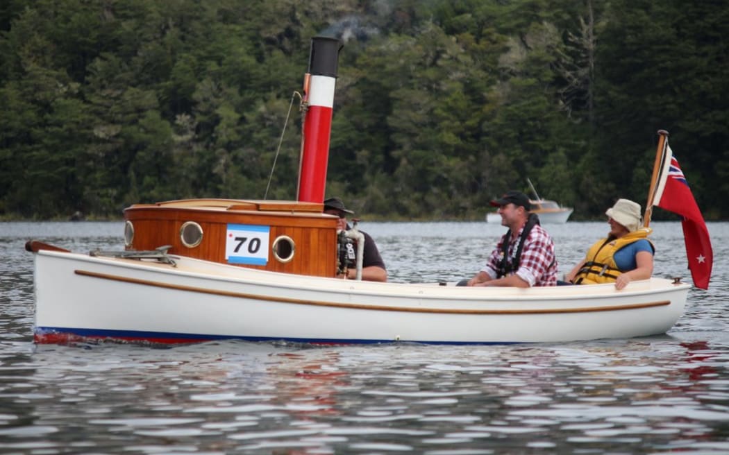 A vintage tugboat with a red chimney. Three people sit in the boat as it glides along a glassy lake, with trees on the shore in the background.