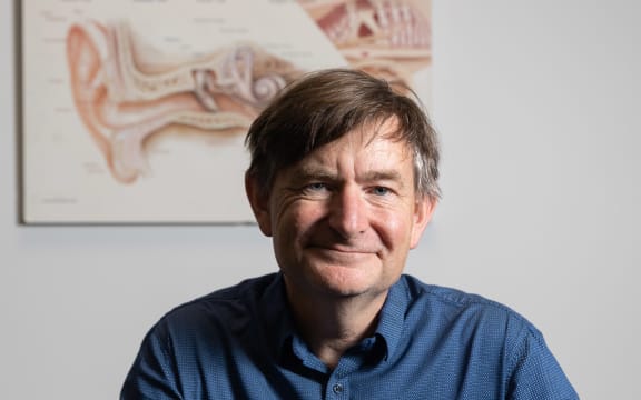 A portrait of Grant. He is in front of a diagram of the anatomy of the ear. He has a wry smile and is wearing a blue shirt.