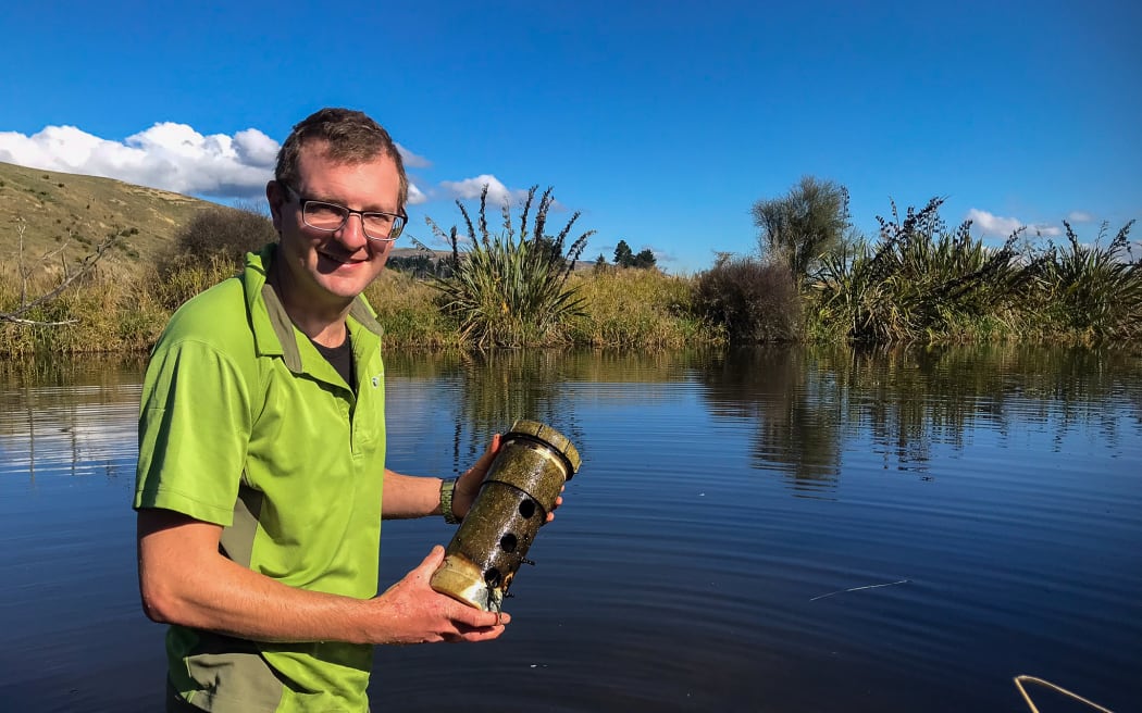 A man stands on the edge of a wetland pool framed by vegetation and long grass. He is holding a pipe-shaped, brown device in his hands, and is posing and smiling at the camera. He is wearing a bright green collared t-shirt and glasses, and the sky is blue and cloudless.