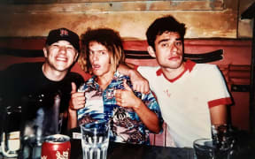 A young DJ StickyFingaz rocking a Hawaiian shirt, and some brown dreadlocks with bleached ends. He's accompanied by two other younger men.
