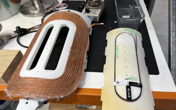 Two pieces of plastic laid on a workbench. On the left hand side large copper coils are wound around in an oblong shape. It is large, and heavy looking. On the right hand side, thin superconducting tape is would around in a slightly elongated oblong shape. Compared to the copper it looks small and light.