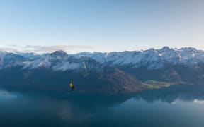 Gavin Alexander Lewis on a highline with the Remarkables in the background. The photo was taken by Will Nelson and is used by RNZ with permission. Will is crouched in the snow on a small ledge at 1400 metres, operating a drone to take the photo.