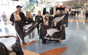 The family of Commonwealth Games' silver medal swimmer Jesse Reynolds wait for him to arrive at Auckland Airport. The six women, one man, and an infant are holding black flags with a silver fern and the words New Zealand.