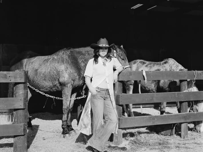 Crystal Chen - a woman wearing flared jeans and a cowboy hat - stands in front of a horse.