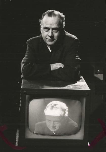 In a black and white photo, Marshall McLuhan, a blading man in a dark suit, rests on a television set with his arms folded.