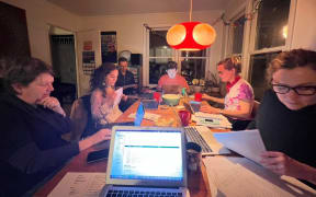 A group of adults gathered in a lounge on a dining table, each with paper work and their laptops out.