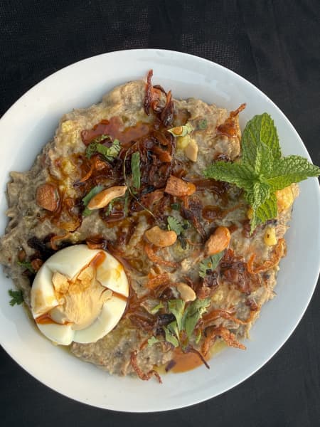 A porridge of Hyderabadi haleem, filled with cooked lentils and meat, topped with a boiled egg, friend onions and herbs.