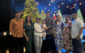 A group photo of seven adults and one child, standing in front of a Christmas tree and smiling at the camera.
