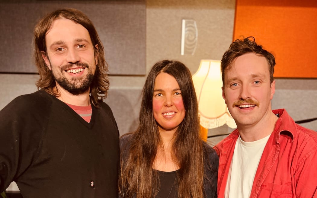 Mel Parsons stands between Josh Logan & Jed Parsons. Mel is holding her guitar. They are all looking towards the camera.