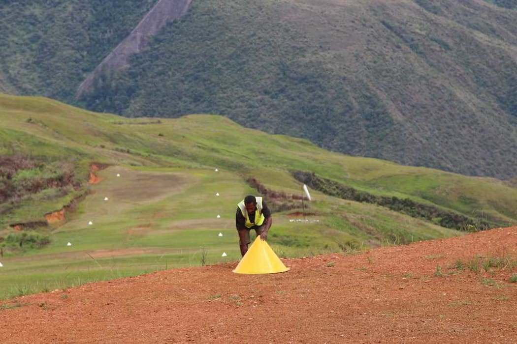 A Papua New Guinea Rural Airstrip Agency Maintenance Officer in Sopu, Goilala places a cone marker. The airstrip and windsocks are visible in the background and mark the taxi way in the distance.