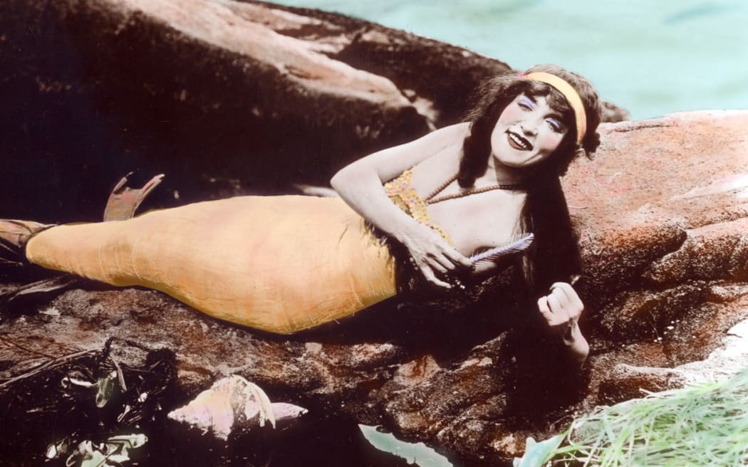 Annette Kellerman in the early 1900s lounging on a rock next to the ocean dressed as a mermaid.