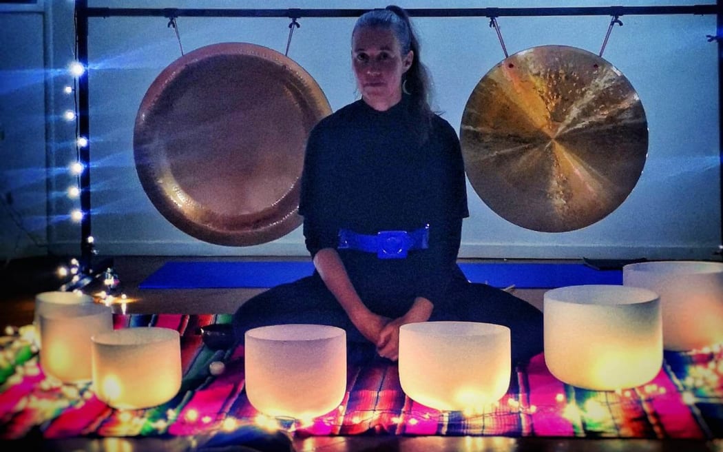 Erika sits cross-legged on a striped mat surrounded by lanterns and fairy lights. There are four gongs hung behind her.