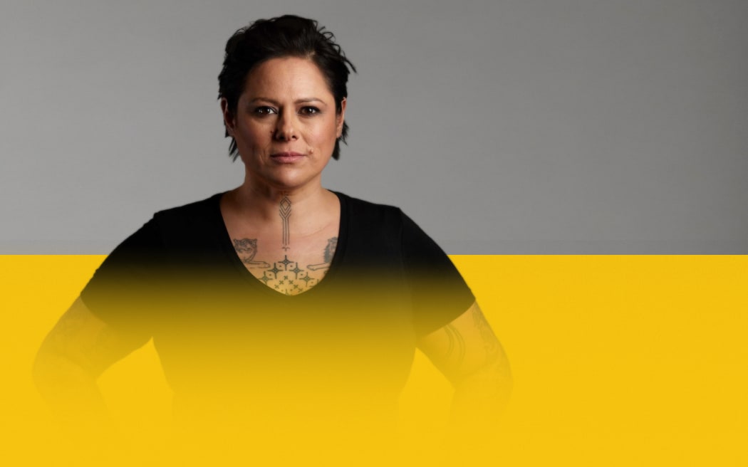 Anika Moa wears a black top with hands on hips, looking straight ahead to camera, on a light grey and yellow background.