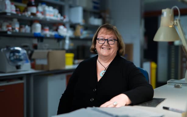 A portrait shot of Laura sitting down at a lab bench. There are shelves of lab equipment visible behind her and a small grey desk lamp in front of her.