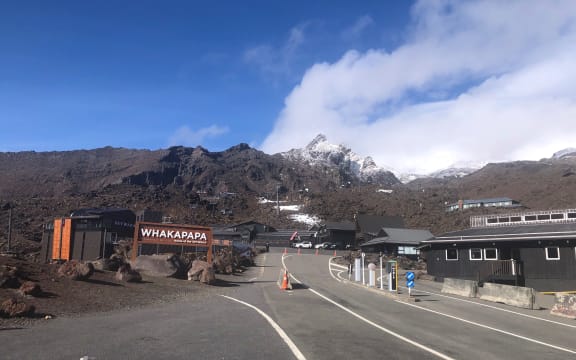 The road into Whakapapa skifield. A wooden sign to the left says "WHAKAPAPA". The sky is a crisp blue. The road disappears up into the snow-capped mountains.
