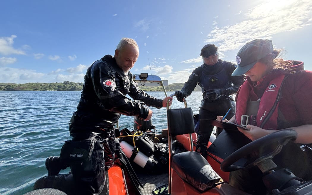 Three people on a small boat; two in diving suits and one writing on a clipboard. The boat is on calm water under a bright sky with scattered clouds.