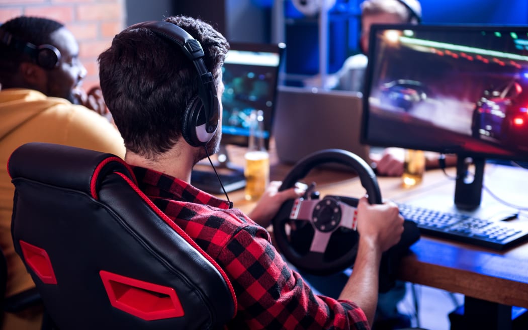 Adult entertainments. Back view of concentrated young man is sitting at desk with friends and playing car racing video game using steering wheel. Males are wearing headphones and resting in background