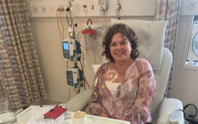 A woman sitting in hospital receiving cancer treatment. A medicines line is embedded into her chest and attached to an IV pump behind her.