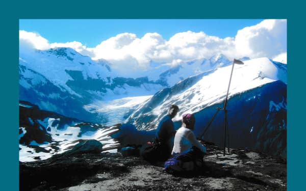 Bluey Green border around a photo taken high up in the mountains. Two people are in the foreground one a flat piece of ground. They are sitting with their tramping packs and appear to be sitting at a trig or other marker. Far below in the valley there is a glacier.