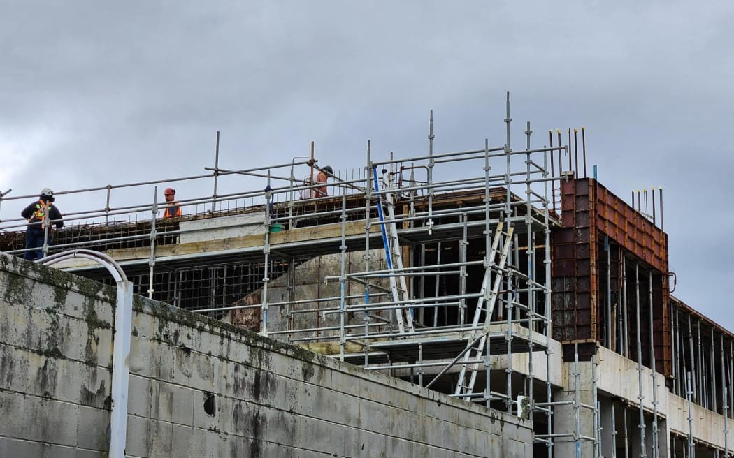 Three construction workers can be seen standing on top of a multi-storey concrete building that is covered by scaffolding.