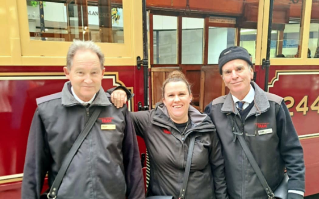 Three people pose in front of a vintage red tram. They are wearing black tram conductor outfits and smiling at the camera.