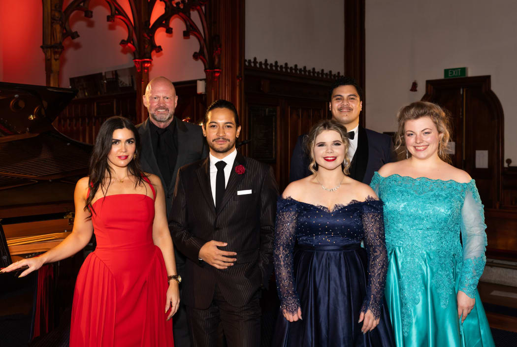 six opera singers on stage in formal ware, smiling at the camera.