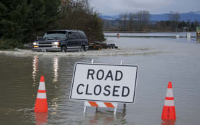 A driver manages to drive through flood waters from the Snohomish River in Snohomish, Washington, on December 11, 2025. Tens of thousands of people were under evacuation orders Thursday in western North America, after days of heavy rain forced rivers to burst their banks. Storms have battered Washington state in the US and British Columbia over the Canadian border for several days, with rivers continuing to rise. (Photo by Jason Redmond / AFP)