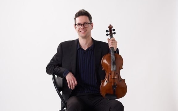 Musician Alexander McFarlane sits on a chair and holds his viola, one end resting on his knee