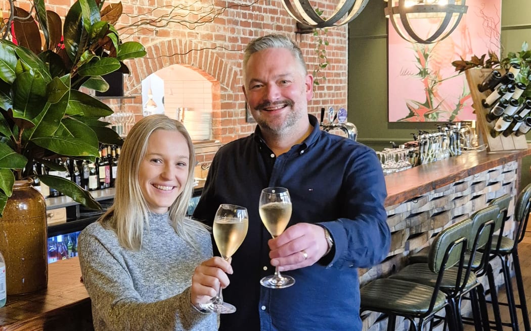 Tim Ogle stands in front of his bar holding a glass of wine. There is a woman stood on his right hand side also holding a glass of wine.