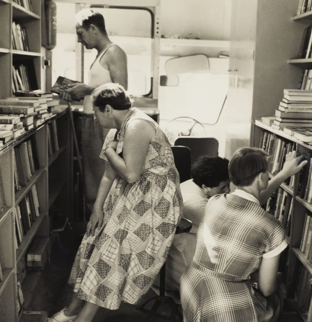 View of three women and a man as seen inside the van that houses the portable library service, shelves of books and a pile of books on a table are visible at left and at right.