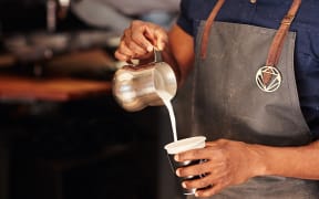 Cropped image of an African barista carefully pouring milk from a stainless steel jug into a takeaway cup in a coffee shop