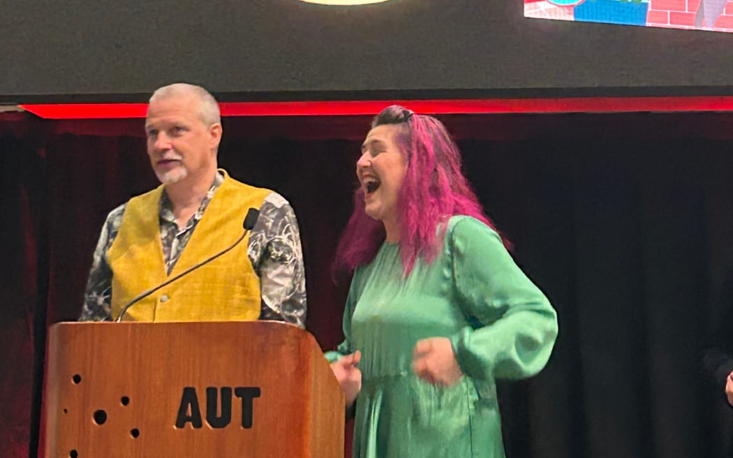 Louise & Gareth Ward are onstage collecting an award at the 2025 'Book industry awards'. Gareth is wearing a waistcoat and looking coy. Louise has her trademark pink hair. She is wearing a green dress and looks very excited. The cover of their book is projected on the stage behind them.