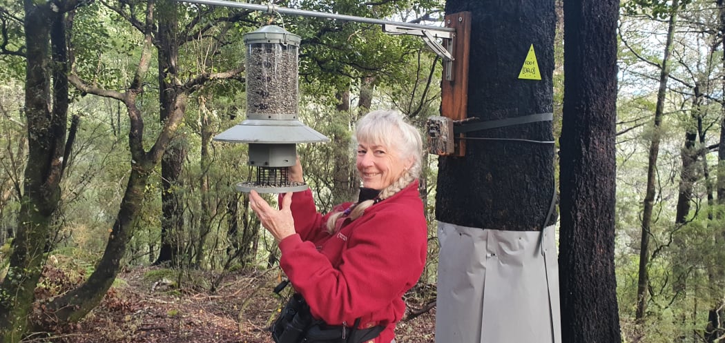 Neroli Amyes is looking towards the camera, smiling, wearing a red jacket. She is holding a large bird feeder which is hanging off a metal cable attached to large pole.