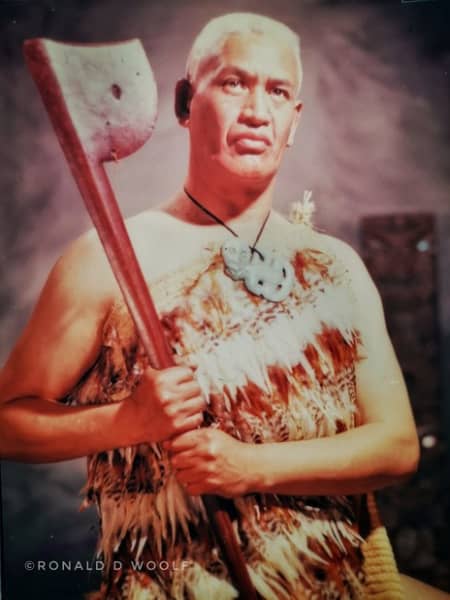 Wiremu (Bill) Kerekere is a grey-haired man in traditional Māori clothing, carrying a large adze.