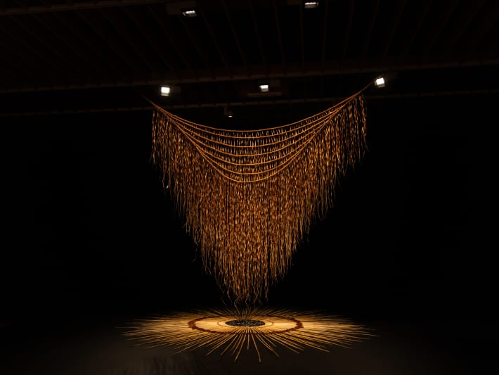 A giant piece of delicate brown weaving is illuminated by lights.