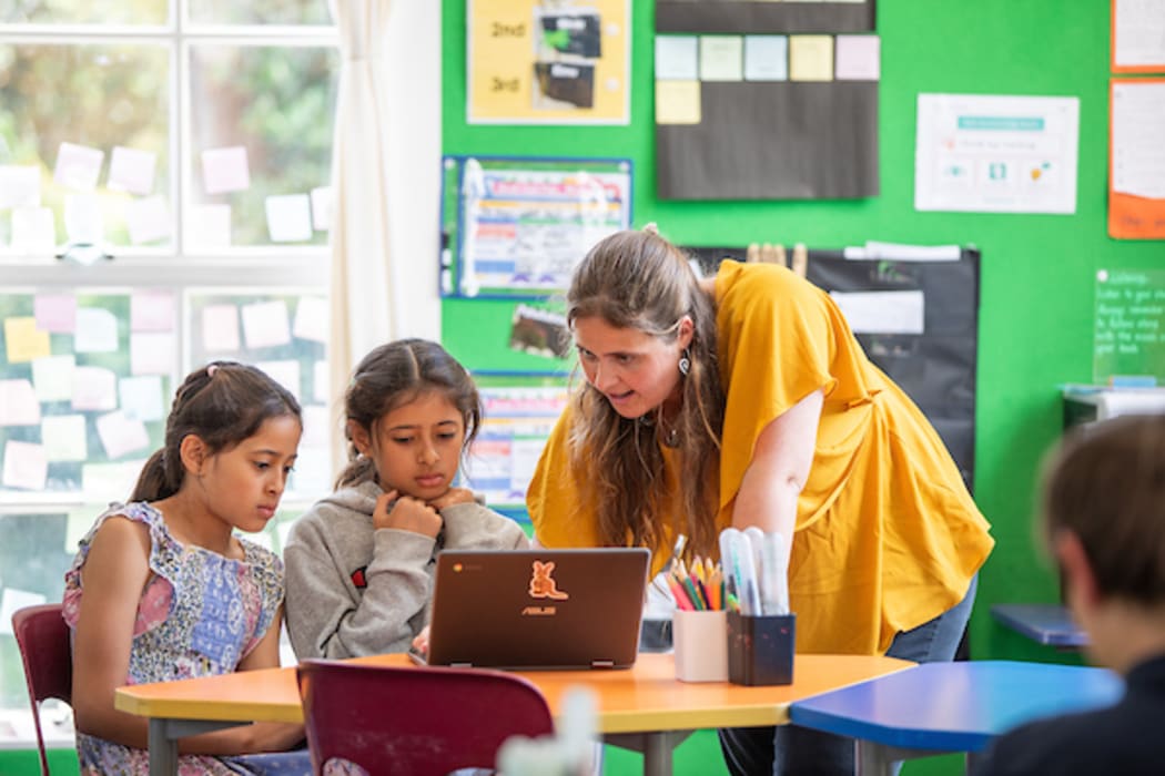 A teacher is leaning over a table in a classroom to help two primary school children who are working together on a laptop.