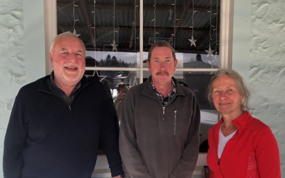 John, Paul and Jill stand on the deck outside a house. They all smile at the camera. The window of the house behind them has starry fairy lights hanging up.