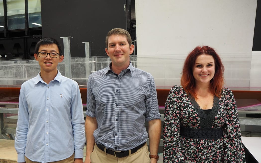 Yaxiong, Colin & Natalia stand in the flume lab with a large perspex rectangle experimental set up behind them.