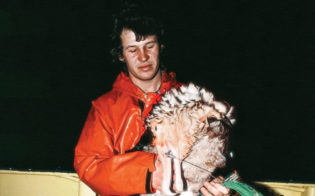 A man wearing a red-orange jacket stands on the deck of a vessel at night. He is looking down at a large dead albatross cradled in his arms, which has a hook protruding from its beak attached to a long coil of green fishing line.