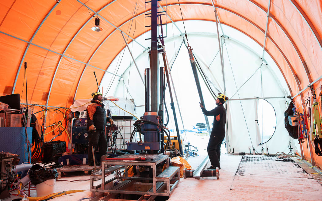 A group of people stand around a complicated piece of metal equipment, inside a large, orange, half-cylindrical tent.