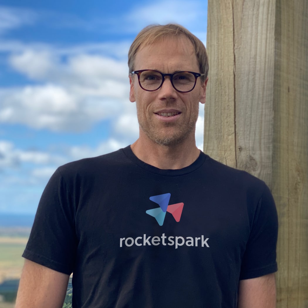 Photo of a man standing in front of blue sky, wearing a t-shirt with "rocketspark" printed on it