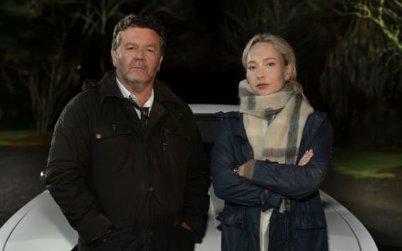 A man and a woman wearing coats give serious looks to the camera while standing in front of a car.