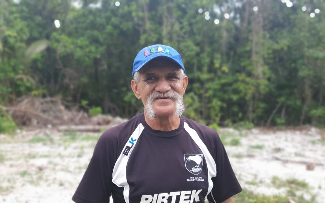 A mid potrait of George Mateariki in front of a forest background, he wears a blue baseball cap and a black and white t-shirt.