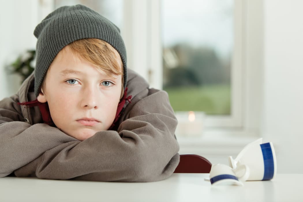 Close Up of Young Teenage Boy Wearing Hoodie and Winter Cap Looking Remorseful While Leaning on Arms in Kitchen with Broken Mug on Table