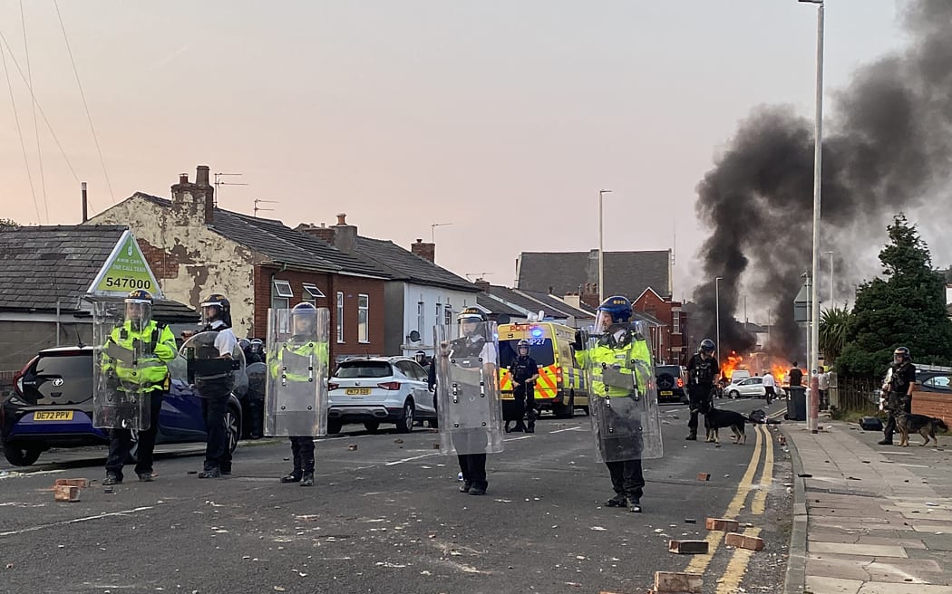 Smoke billows from a fire started by protesters as riot police stand guard after disturbances near the Southport Islamic Society Mosque in Southport, northwest England, on July 30, 2024, a day after a deadly child knife attack. Violent clashes broke out in the northern England town where a knife attack claimed the lives of three children, with around 100 protesters lighting fires and battling police. A 17-year-old male suspect from a nearby village arrested shortly after the incident remained in custody, police added, as they warned against speculating about his identity or details of the investigation. (Photo by Roland LLOYD PARRY / AFP)