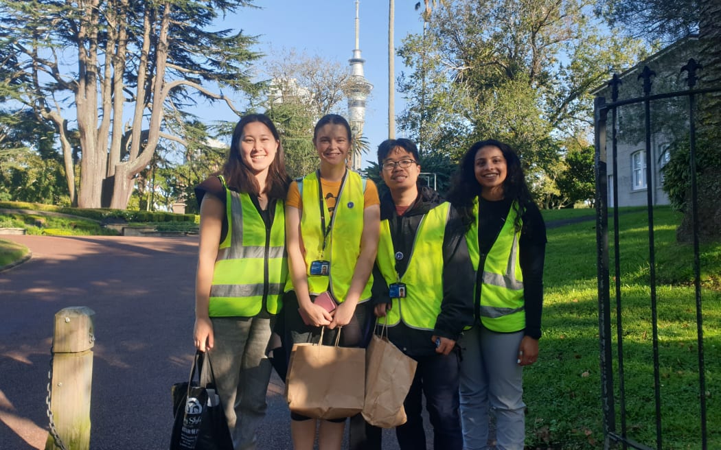 Four people in high-vis vests stand at the entrance to a park on a sunny morning. The Sky Tower is visible in the background. The people are smiling.