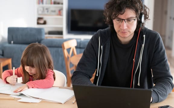 Young father working from home with little daughter drawing during covid-19 lockdown. Child drawing next to dad who is smartworking with laptop and headphones for social isolation. Front view.