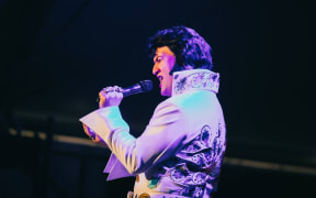 Taurean Kenny Mill holds a microphone as he performs on stage in full Elvis Presley attire.